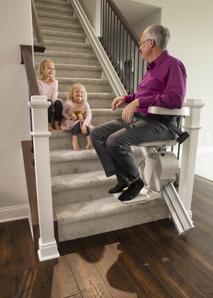 A man riding a Bruno Elan stairlift with his grandchildren on the steps