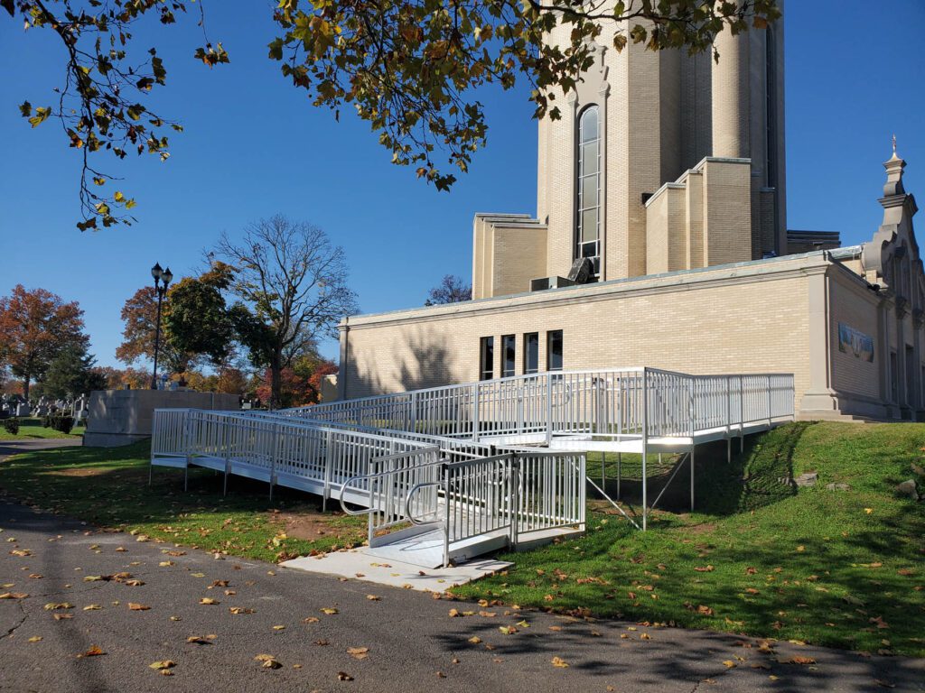 A long aluminum wheelchair ramp switching back along the side of a stone building, with handrails on both sides