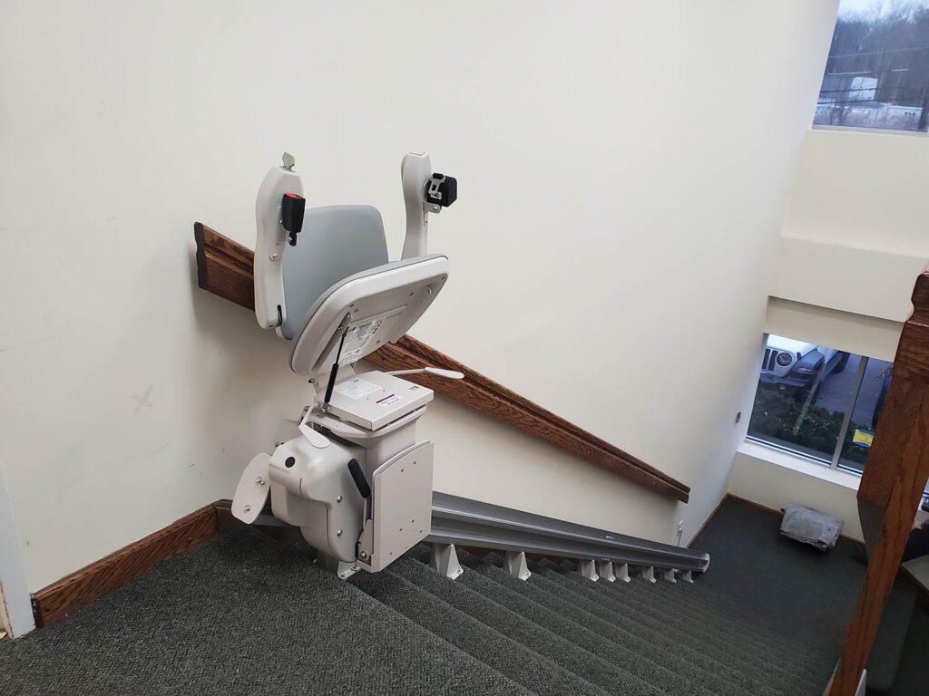 A straight stairlift folded up at the top of a carpeted office stairwell, the rail running down the treads along the handrail