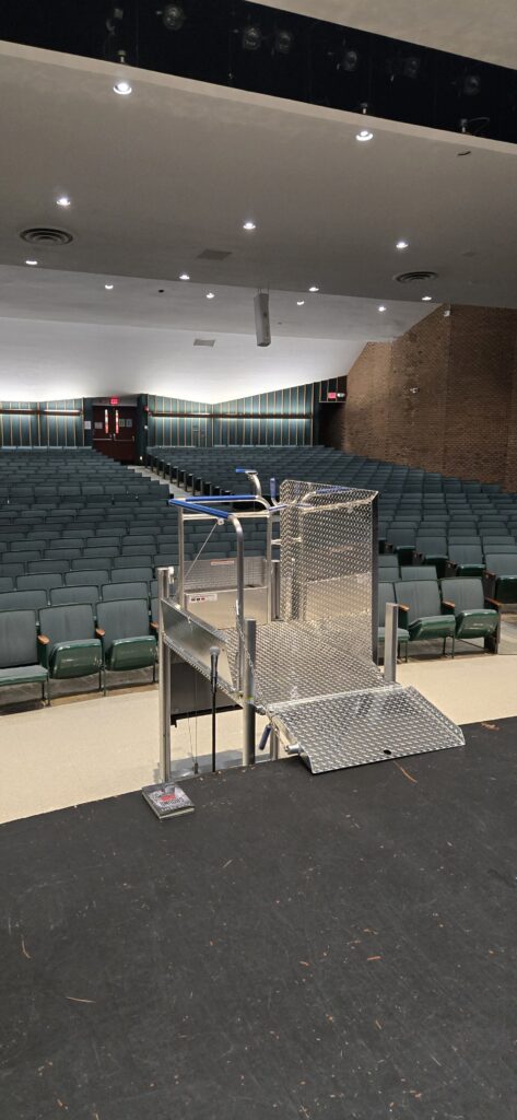 A Mobilift CX portable wheelchair lift at the foot of an auditorium stage, its bridge ramp down, with rows of green seats behind