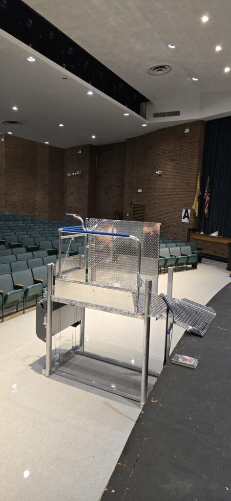 A Mobilift CX portable wheelchair lift set up in front of an auditorium stage, seen from the side