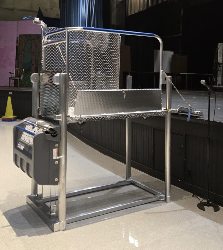 A Mobilift CX portable wheelchair lift beside an auditorium stage, its platform raised to stage height